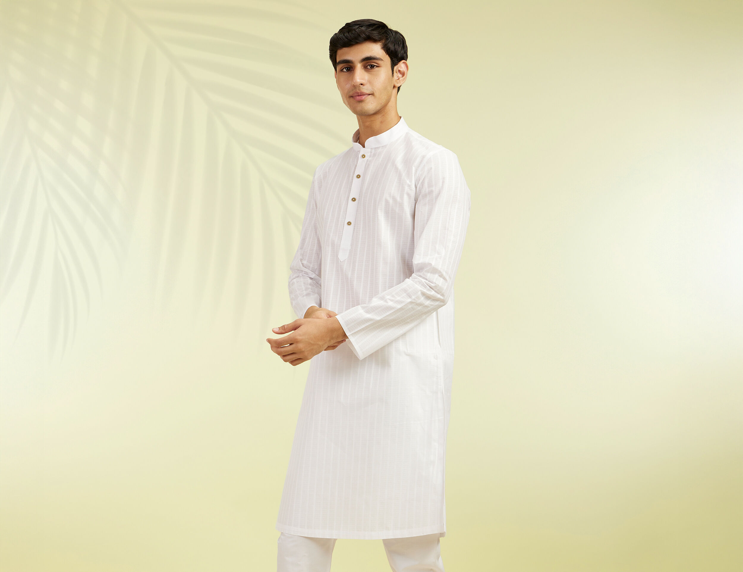Man in an elegant white kurta-pajama with button detailing, styled against a soft beige background with subtle palm leaf shadows, showcasing a refined Onam festive look.